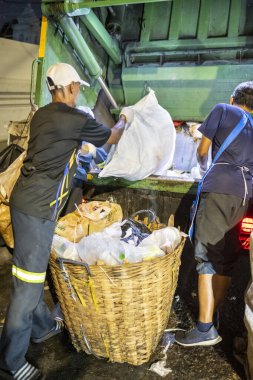 Bangkok, Tayland-26 Nisan 2025: Belediye çöp toplayıcıları, yerel dükkanlar ve evlerin çöpleriyle dolu bambu sepetlerini, bekleyen bir çöp arabasının arkasındaki makineler tarafından ezilmek üzere atıyorlar.