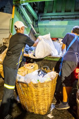 Bangkok, Tayland-26 Nisan 2025: Belediye çöp toplayıcıları, yerel dükkanlar ve evlerin çöpleriyle dolu bambu sepetlerini, bekleyen bir çöp arabasının arkasındaki makineler tarafından ezilmek üzere atıyorlar.