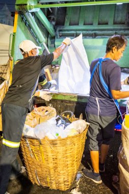 Bangkok, Tayland-26 Nisan 2025: Belediye çöp toplayıcıları, yerel dükkanlar ve evlerin çöpleriyle dolu bambu sepetlerini, bekleyen bir çöp arabasının arkasındaki makineler tarafından ezilmek üzere atıyorlar.