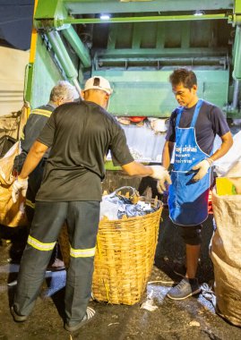Bangkok, Tayland-26 Nisan 2025: Belediye çöp toplayıcıları, yerel dükkanlar ve evlerin çöpleriyle dolu bambu sepetlerini, bekleyen bir çöp arabasının arkasındaki makineler tarafından ezilmek üzere atıyorlar.