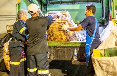 Bangkok, Tayland-26 Nisan 2025: Belediye çöp toplayıcıları, yerel dükkanlar ve evlerin çöpleriyle dolu bambu sepetlerini, bekleyen bir çöp arabasının arkasındaki makineler tarafından ezilmek üzere atıyorlar.