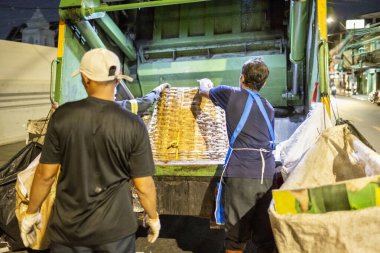 Bangkok, Tayland-26 Nisan 2025: Belediye çöp toplayıcıları, yerel dükkanlar ve evlerin çöpleriyle dolu bambu sepetlerini, bekleyen bir çöp arabasının arkasındaki makineler tarafından ezilmek üzere atıyorlar.