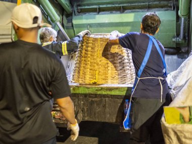 Bangkok, Tayland-26 Nisan 2025: Belediye çöp toplayıcıları, yerel dükkanlar ve evlerin çöpleriyle dolu bambu sepetlerini, bekleyen bir çöp arabasının arkasındaki makineler tarafından ezilmek üzere atıyorlar.