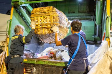 Bangkok, Tayland-26 Nisan 2025: Belediye çöp toplayıcıları, yerel dükkanlar ve evlerin çöpleriyle dolu bambu sepetlerini, bekleyen bir çöp arabasının arkasındaki makineler tarafından ezilmek üzere atıyorlar.