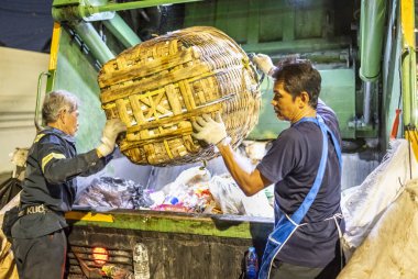 Bangkok, Tayland-26 Nisan 2025: Belediye çöp toplayıcıları, yerel dükkanlar ve evlerin çöpleriyle dolu bambu sepetlerini, bekleyen bir çöp arabasının arkasındaki makineler tarafından ezilmek üzere atıyorlar.