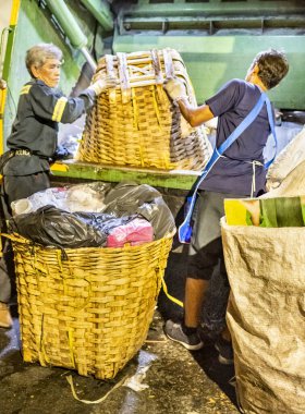 Bangkok, Tayland-26 Nisan 2025: Belediye çöp toplayıcıları, yerel dükkanlar ve evlerin çöpleriyle dolu bambu sepetlerini, bekleyen bir çöp arabasının arkasındaki makineler tarafından ezilmek üzere atıyorlar.