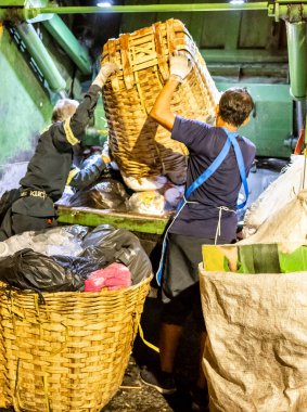 Bangkok, Tayland-26 Nisan 2025: Belediye çöp toplayıcıları, yerel dükkanlar ve evlerin çöpleriyle dolu bambu sepetlerini, bekleyen bir çöp arabasının arkasındaki makineler tarafından ezilmek üzere atıyorlar.