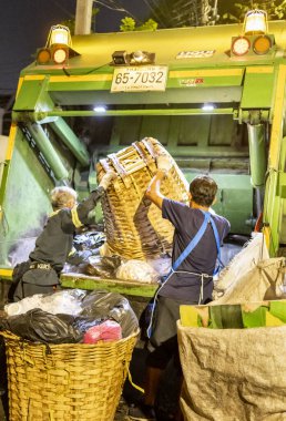 Bangkok, Tayland-26 Nisan 2025: Belediye çöp toplayıcıları, yerel dükkanlar ve evlerin çöpleriyle dolu bambu sepetlerini, bekleyen bir çöp arabasının arkasındaki makineler tarafından ezilmek üzere atıyorlar.