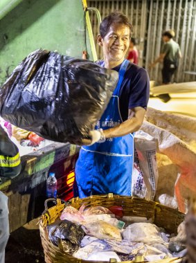 Bangkok, Tayland-26 Nisan 2025: Belediye çöp toplayıcıları, yerel dükkanlar ve evlerin çöpleriyle dolu bambu sepetlerini, bekleyen bir çöp arabasının arkasındaki makineler tarafından ezilmek üzere atıyorlar.