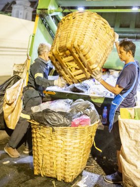Bangkok, Tayland-26 Nisan 2025: Belediye çöp toplayıcıları, yerel dükkanlar ve evlerin çöpleriyle dolu bambu sepetlerini, bekleyen bir çöp arabasının arkasındaki makineler tarafından ezilmek üzere atıyorlar.