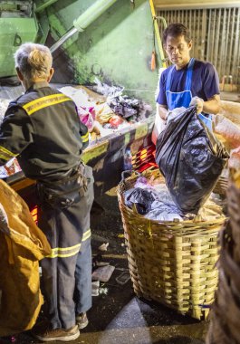 Bangkok, Tayland-26 Nisan 2025: Belediye çöp toplayıcıları, yerel dükkanlar ve evlerin çöpleriyle dolu bambu sepetlerini, bekleyen bir çöp arabasının arkasındaki makineler tarafından ezilmek üzere atıyorlar.