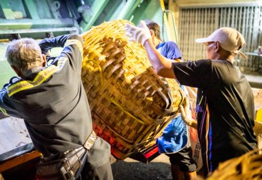 Bangkok, Tayland-26 Nisan 2025: Belediye çöp toplayıcıları, yerel dükkanlar ve evlerin çöpleriyle dolu bambu sepetlerini, bekleyen bir çöp arabasının arkasındaki makineler tarafından ezilmek üzere atıyorlar.