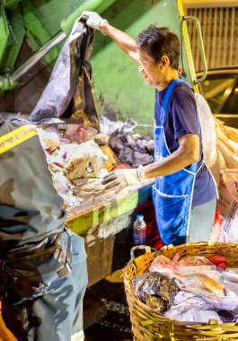Bangkok, Tayland-26 Nisan 2025: Belediye çöp toplayıcıları, yerel dükkanlar ve evlerin çöpleriyle dolu bambu sepetlerini, bekleyen bir çöp arabasının arkasındaki makineler tarafından ezilmek üzere atıyorlar.