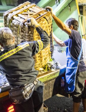 Bangkok, Tayland-26 Nisan 2025: Belediye çöp toplayıcıları, yerel dükkanlar ve evlerin çöpleriyle dolu bambu sepetlerini, bekleyen bir çöp arabasının arkasındaki makineler tarafından ezilmek üzere atıyorlar.