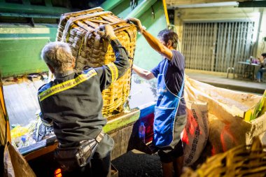 Bangkok, Tayland-26 Nisan 2025: Belediye çöp toplayıcıları, yerel dükkanlar ve evlerin çöpleriyle dolu bambu sepetlerini, bekleyen bir çöp arabasının arkasındaki makineler tarafından ezilmek üzere atıyorlar.