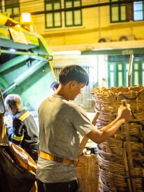 Bangkok, Tayland-26 Nisan 2025: Belediye çöp toplayıcıları, yerel dükkanlar ve evlerin çöpleriyle dolu bambu sepetlerini, bekleyen bir çöp arabasının arkasındaki makineler tarafından ezilmek üzere atıyorlar.