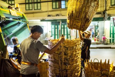Bangkok, Tayland-26 Nisan 2025: Belediye çöp toplayıcıları, yerel dükkanlar ve evlerin çöpleriyle dolu bambu sepetlerini, bekleyen bir çöp arabasının arkasındaki makineler tarafından ezilmek üzere atıyorlar.
