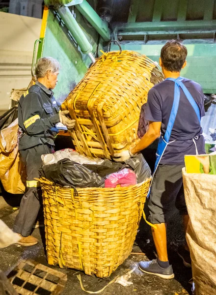 Bangkok, Tayland-26 Nisan 2025: Belediye çöp toplayıcıları, yerel dükkanlar ve evlerin çöpleriyle dolu bambu sepetlerini, bekleyen bir çöp arabasının arkasındaki makineler tarafından ezilmek üzere atıyorlar.