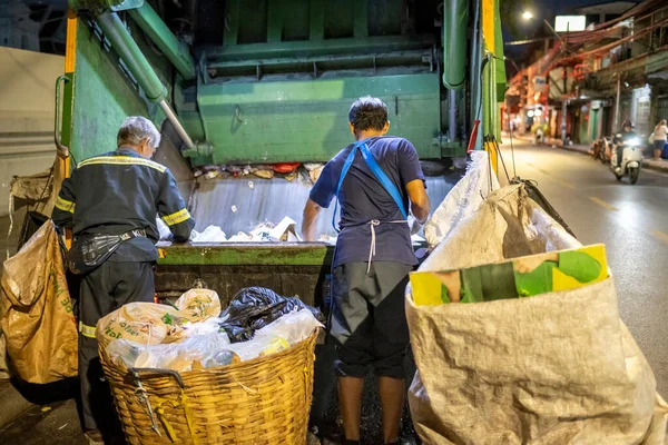 Bangkok, Tayland-26 Nisan 2025: Belediye çöp toplayıcıları, yerel dükkanlar ve evlerin çöpleriyle dolu bambu sepetlerini, bekleyen bir çöp arabasının arkasındaki makineler tarafından ezilmek üzere atıyorlar.