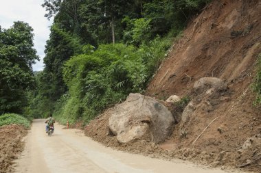Tehlikeli heyelan, uzak dağlardaki kırsal toprak yolu tıkayan büyük kayalar. Motosikletli kişi tehlikeli yollarda geziniyor.