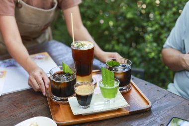 Barista açık bir kafede çeşitli buzlu kahve ve karışık içecekler servis ediyor. Müşteri menüden ahşap masaya içecek koyuyor.