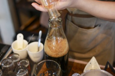 Profesyonel barmen cam şişeye şurup dolduruyor, ustalıkla özel kahve içeceği yapıyor. Konforlu kafe içindeki bu odaklanmış içecek hazırlığı zanaat sürecini gösteriyor