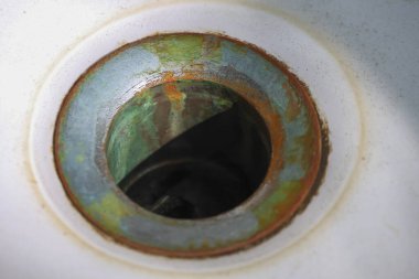 Metal deliğin etrafındaki pas ve aşınmayı gösteren eski, kirli bir lavabo giderinin makro görüntüsü. Bu bakımsız banyo armatürü hijyenik görünmüyor ve tamir edilmesi gerekiyor.