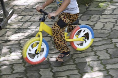 Leopar desenli pantolonlu, sarı dengeli bisiklet sürmeyi öğrenen bir çocuk. Asfaltlı arka plandaki açık hava aktivitesi oyun ve çocukluk gelişiminin mutlu anını gösteriyor