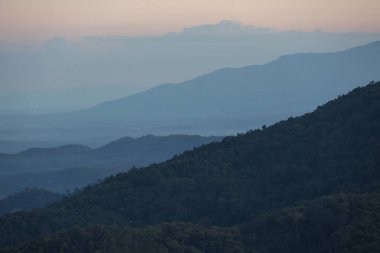 Alacakaranlıkta sakin dağ manzarası. Mavi tepeler ve vadilerden oluşan katmanlar, yumuşak gökyüzünün altında bulanık bir mesafeye çekilip, doğanın huzurlu ve görkemli manzarasını yaratıyor.