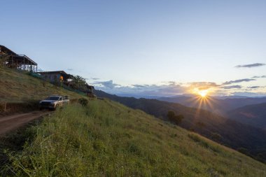 Uzak dağ manzarasında, toprak yolda kamyoneti olan huzurlu bir gün doğumu. Ahşap kulübe yeşil tepede oturuyor manzaralı vadi ve sabah güneşine bakan