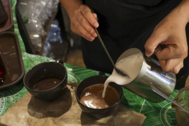 Odaklanmış barista buğulanmış süt köpüğünü bir fincan kahveye döküyor. Yetenekli eller sıcak latte yapıyor. Sanat içeceği, el işi ve kafe hazırlığı.