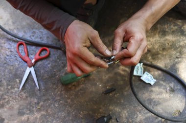 Odaklanmış işçi elleri, beton zeminde metal klemple siyah kabloyu dikkatlice onarıyor. Bu gayretli el emeği, yoğun konsantrasyonla bağlantının düzeltildiğini gösteriyor