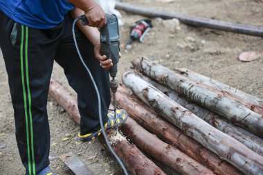 İnşaat alanında elektrikli matkapla ahşap kütüğe delik açmaya odaklanmış bir adam. Bu işçi aktivitesi el emeği, ahşap işçiliği ve inşaat içeriyor.