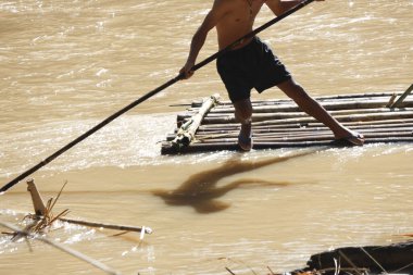 Bambu salına odaklanmış Asyalı bir adam çamurlu nehirde uzun bir direk kullanıyor. Bu geleneksel su seyahati metodu çalışkan bir yaşam tarzı gösteriyor.