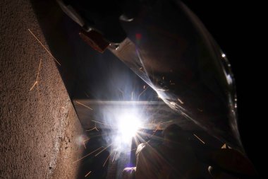 Parlak ışık ve uçan kıvılcımla yoğun kaynak işlemi. Kaynakçı karanlık atölyede metal üretiminde çalışıyor çelik parçalarını endüstriyel beceri ve hassasiyetle birleştiriyor.