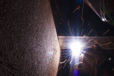 Endüstriyel kaynaklara yakın mesafede parlak ışık yayları ve sıcak uçan kıvılcımlar görünüyor. Yetenekli işçiler karanlık atölyede yoğun odaklanma ve hassasiyetle metal üretiyorlar.