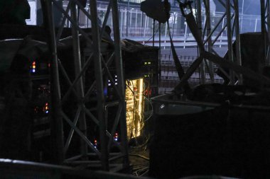 Karanlık endüstriyel ortamda sahne arkasında gizemli parlayan elektrikli ekipmanlar. Metal yapının içinde ışık olan fütüristik güç kaynağı gergin bir ortam yaratıyor.