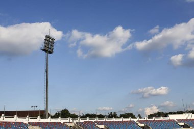 Stadyum ışıkları beyaz bulutlu parlak mavi gökyüzüne karşı dimdik ayakta duruyor. Bu barışçıl açık hava spor sahnesi büyük maç veya etkinlik öncesi sessizlik hissi uyandırıyor.