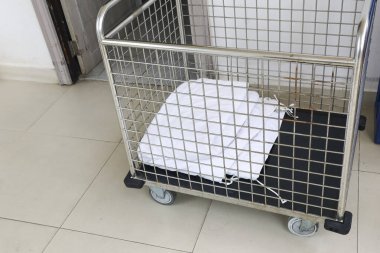 Boş bir endüstriyel metal çamaşır sepetinin içinde yalnız beyaz giysi parçası. Servis arabası odada bekliyor. Temizlik işi için yalnız ve sessiz bir ortam yaratıyor.