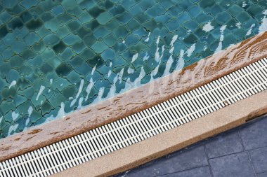 Yüzme havuzu kenarından akan ferahlatıcı mavi suyu taşan bir ızgaraya yakın tut. Temiz su, mozaik fayans ve drenaj sistemli sakin havuz kenarı manzarası