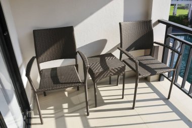 Güneşli otel balkonunda sandalyeli ve masalı bir açık hava sahnesi. Bu boş mobilyalar sabah güneşinde sessiz bir huzur ve rahatlık anına davetiye çıkarır.