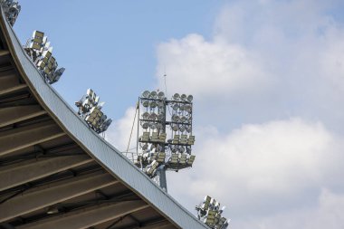 Mavi gökyüzüne karşı geniş arenanın metal çatı yapısında stadyum ışığı kümesi. Güçlü açık hava aydınlatması için profesyonel spor ekipmanları