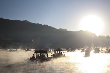 Güneşin sabah sisli gölün üzerinde, teknelerle doğuşu. Barışçıl turdaki insanlar sakin suların ve sıcak, sakinleştirici güneş ışığıyla güzel puslu manzaranın tadını çıkarıyorlar.