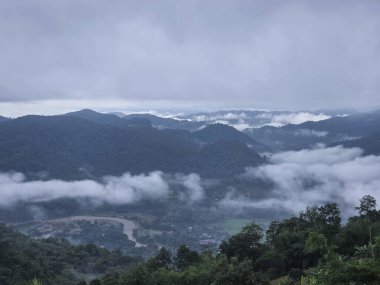 Bulutlu ve bulutlu bir günde sakin ve görkemli bir dağ manzarası. Bu yeşil arazi, nehir ve alçak sisli vadiyi barındırıyor. Huzursuz ve karamsar bir manzara yaratıyor.