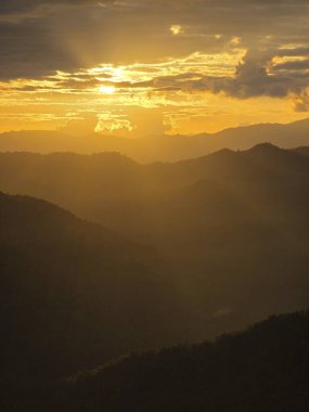 Dağ siluetinin üzerinde güzel altın gün batımı. Doğanın huzurlu manzarası dramatik gökyüzü, bulutlar ve sıcak ışık huzmeleri vadide parlıyor.