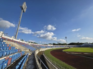 Güneşli bir günde mavi koltuklu boş bir atletizm stadyumunun geniş açılı manzarası. Bir sonraki büyük yarışma için barışçıl bir beklenti hissi
