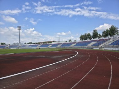 Spor müsabakası için kırmızı koşu pisti ve mavi koltuklu boş bir stadyum. Bulutlu mavi gökyüzü altında atletik arena sessiz bir beklenti hissi veriyor.