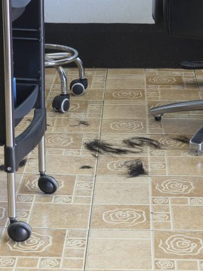 Dağınık bir salon zemininde koyu renk saç, ortalığı karıştırıyor. Temizlenmesi gereken profesyonel bir berber işi. Moda sandalye ve el arabasının yanındaki fayanslı zeminde saç budama manzarası