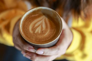 Güzel latte sanatıyla sıcak, sıcak bir fincan kahve içen bir kadının elini yakın çekim. Sabahları kafede sıcak bir içecekle rahatlatıcı anın tadını çıkarıyorum.
