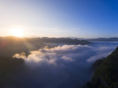 Gün doğumunda sakin bir dağ manzarası. Altın güneş bulutların ve sislerin üzerinde parlıyor. Tropikal vadide huzurlu ve görkemli bir sabah manzarası yaratıyor.